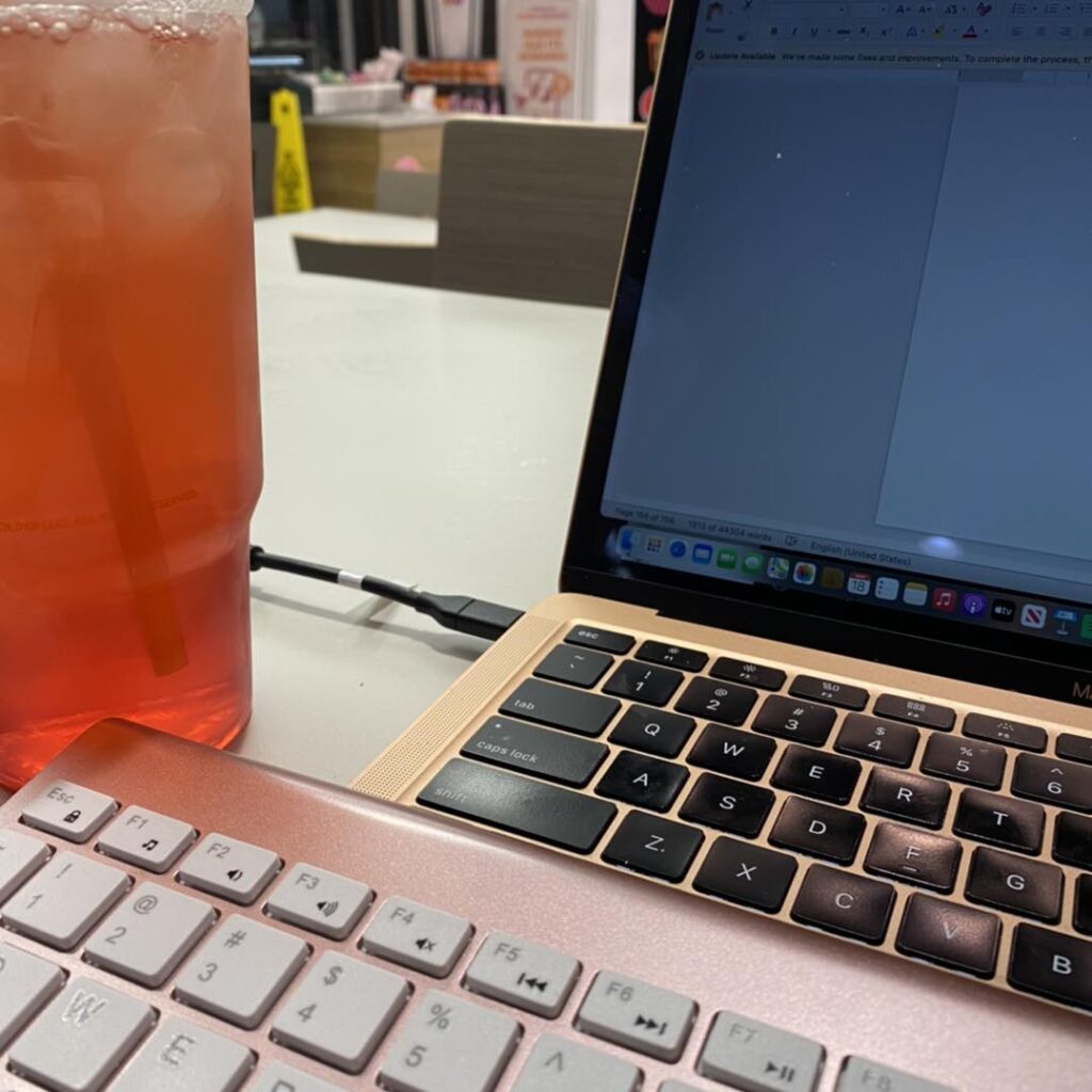 A computer and keyboard in the foreground with a tall pink drink and a cafe in the background